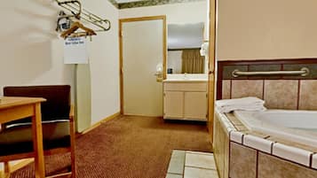 Habitación de hotel con cama, un escritorio pequeño, una silla, baño con lavabo y espejo, y una puerta que da a otra habitación.