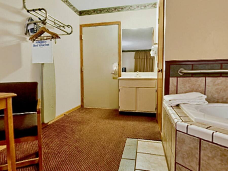Habitación de hotel con cama, un escritorio pequeño, una silla, baño con lavabo y espejo, y una puerta que da a otra habitación.