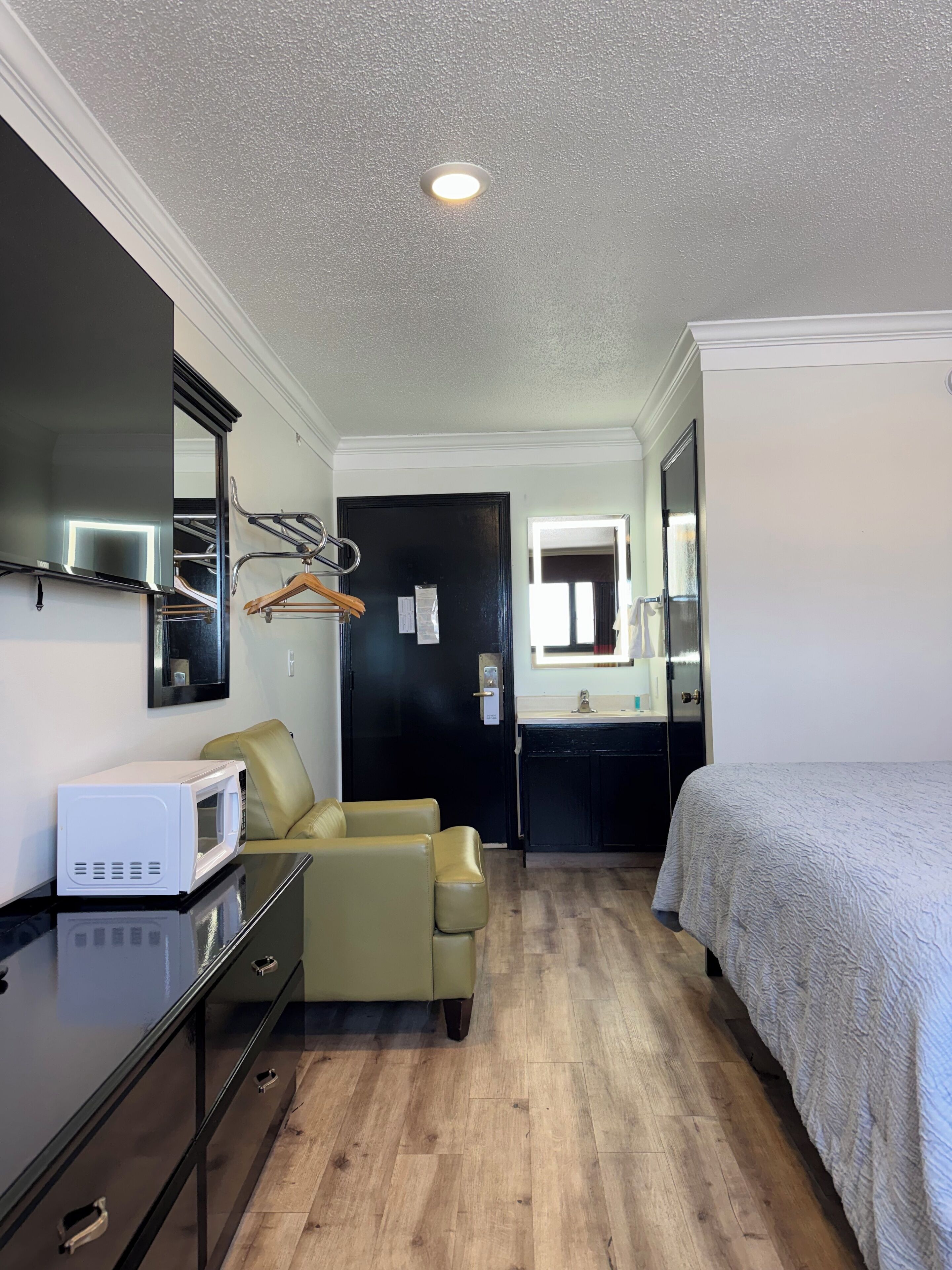 Ein kompaktes Hotelzimmer mit einem Bett, einem kleinen Schreibtisch mit einer Mikrowelle, einem Stuhl, einem Spiegel und einer Tür zum Badezimmer.