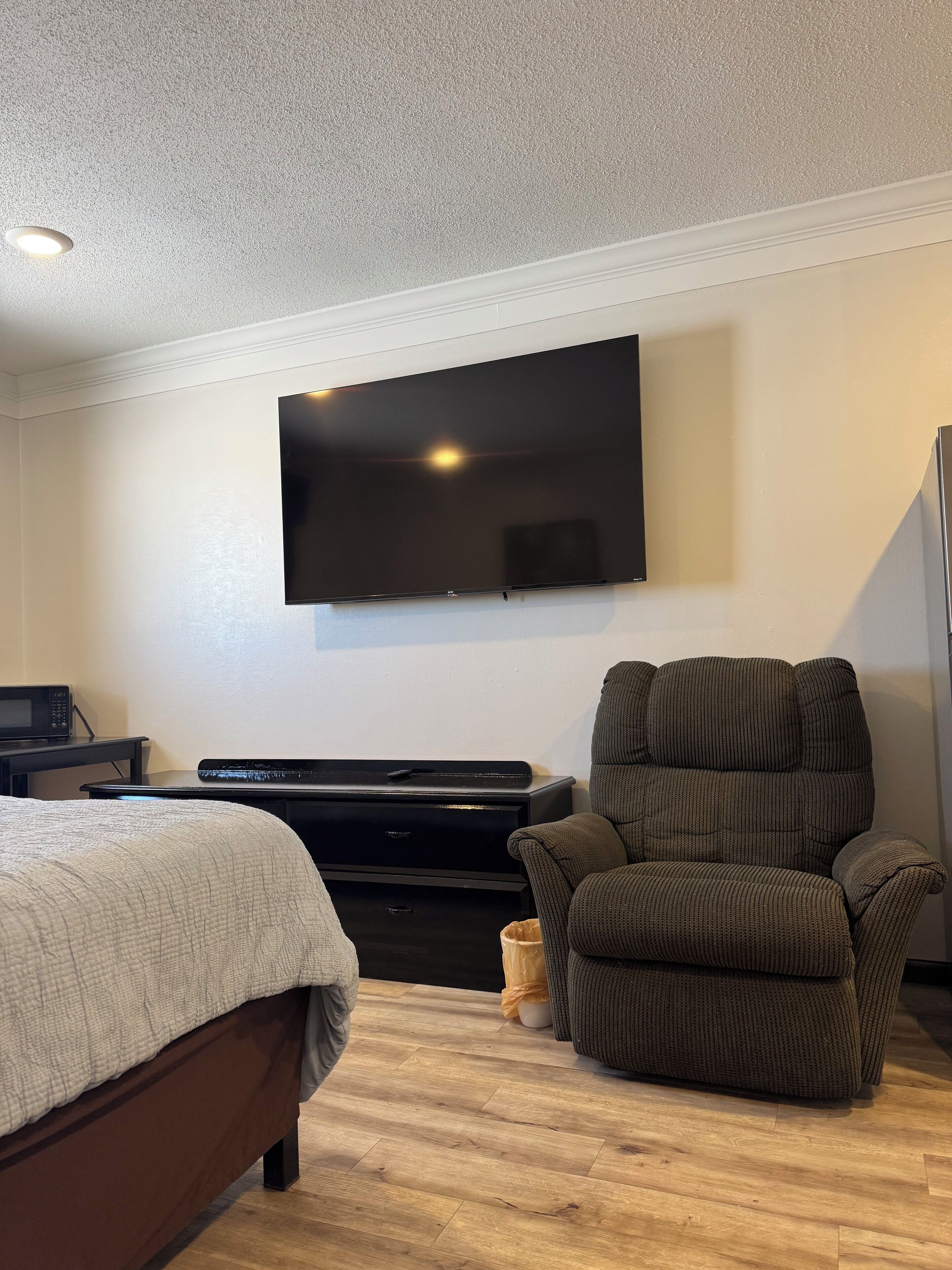 A room with a bed, a dark-colored armchair, a flat-screen TV mounted on the wall, a small table with a microwave, and a wooden floor.