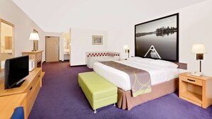 A hotel room with a bed, a TV, a desk, a chair, a green ottoman, a nightstand, a lamp, and a painting on the wall. - Super 8 by Wyndham St. James (Saint James)