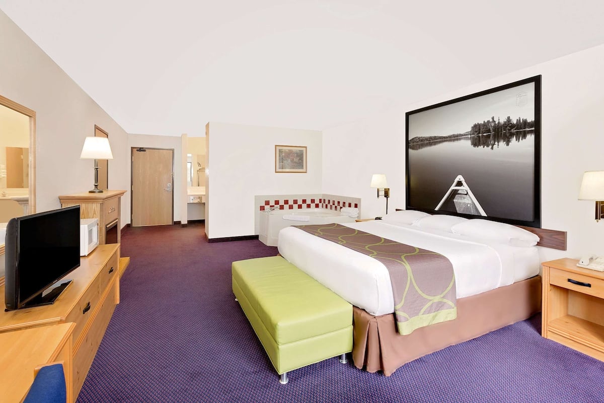 Habitación de hotel con cama, televisor, escritorio, silla, reposapiés verde, mesita de noche, lámpara y un cuadro en la pared.
