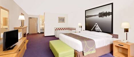 A hotel room with a bed, a TV, a desk, a chair, a green ottoman, a nightstand, a lamp, and a painting on the wall.