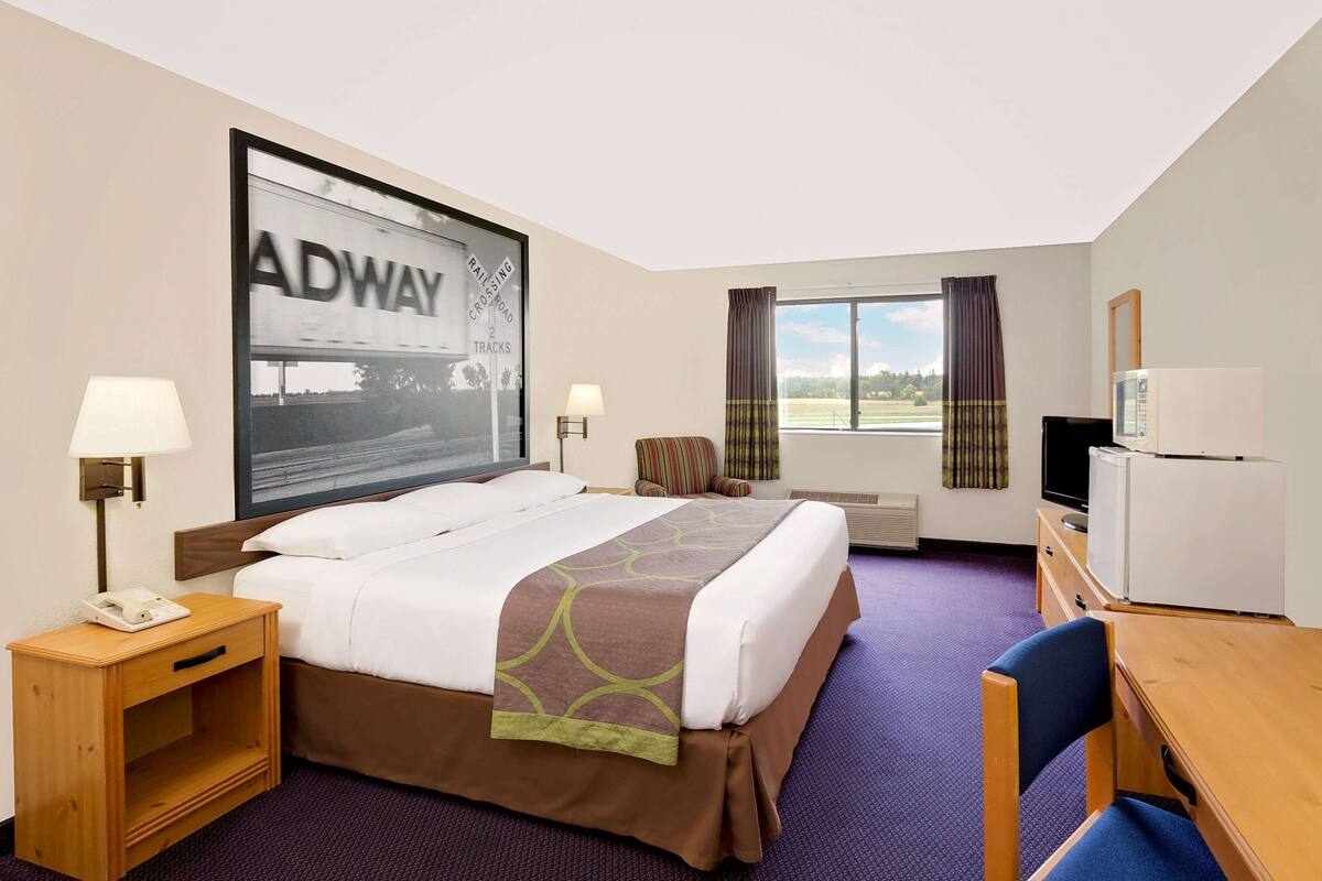 Habitación de hotel con una cama grande, un escritorio, una silla, un televisor, un refrigerador, una lámpara, una mesita de noche y una ventana con cortinas.