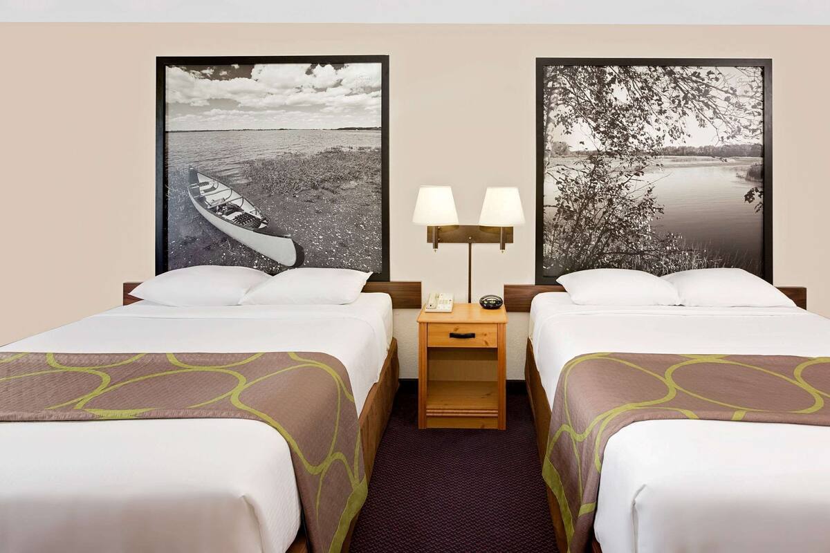 Habitación de hotel con dos camas, una mesita de noche con lámpara y dos cuadros enmarcados en la pared.