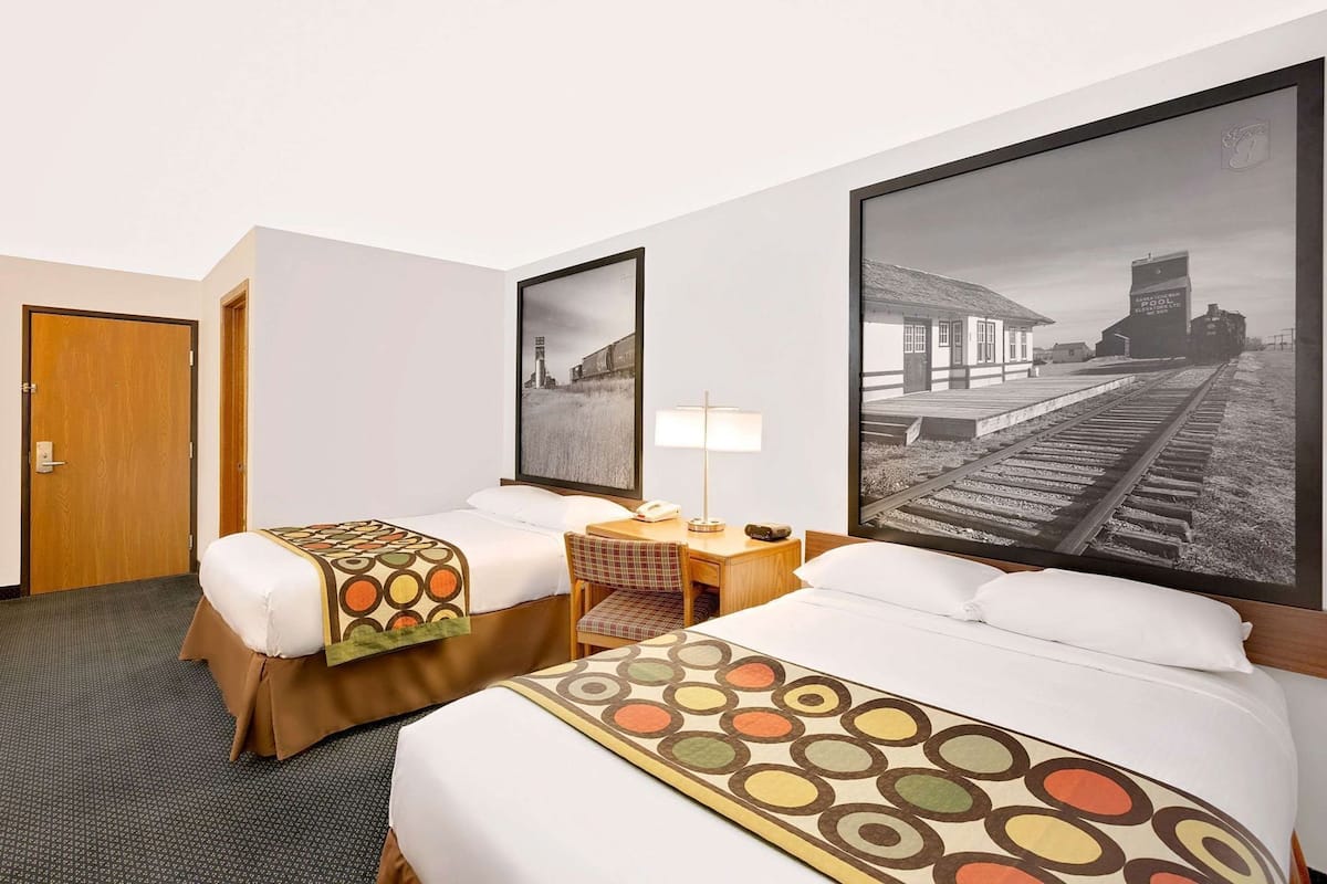 Habitación de hotel con dos camas, un escritorio, una silla, una lámpara y un cuadro grande con una estación de tren.