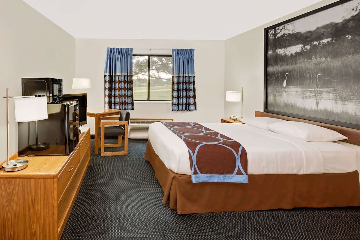Habitación de hotel con cama, escritorio, silla, televisor y un ventanal grande con cortinas.