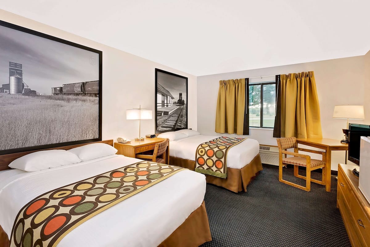 Habitación de hotel con dos camas, un escritorio, una silla, un televisor y dos cuadros grandes en la pared.