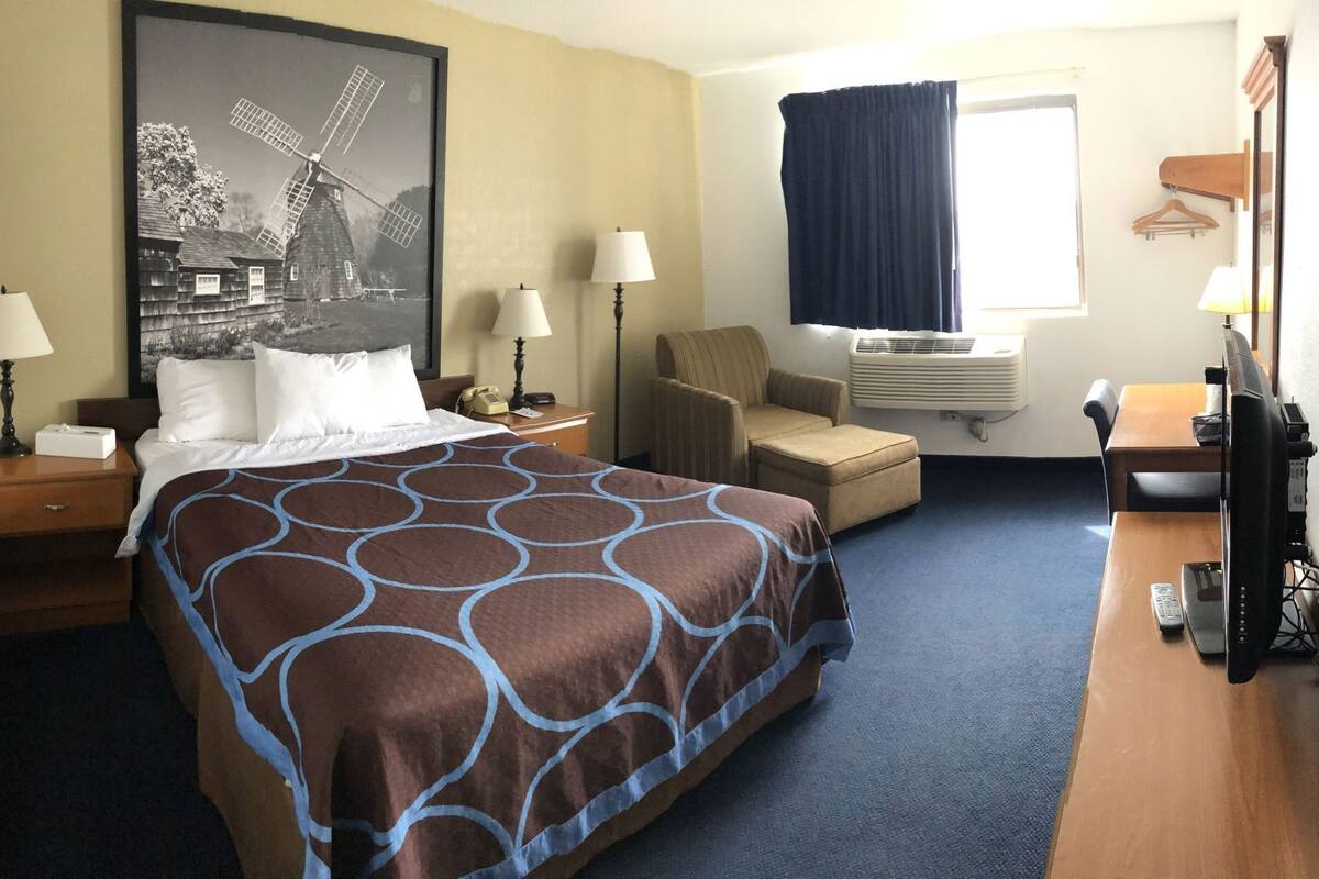 A hotel room with a bed, two bedside tables, a chair, a desk, a television, and an air conditioning unit.