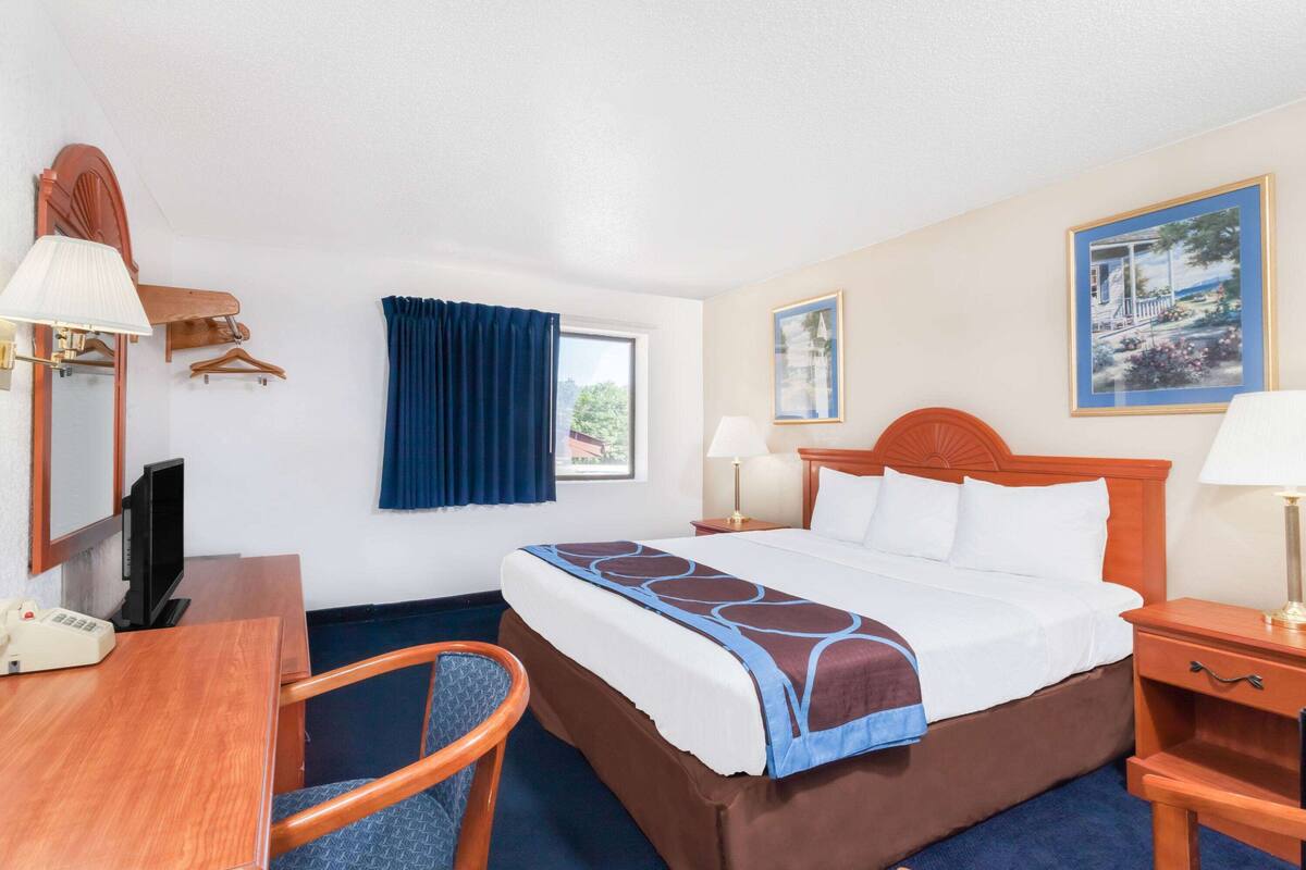 A hotel room with a bed, bedside tables, a desk with a telephone, a chair, a television, and a window with curtains.