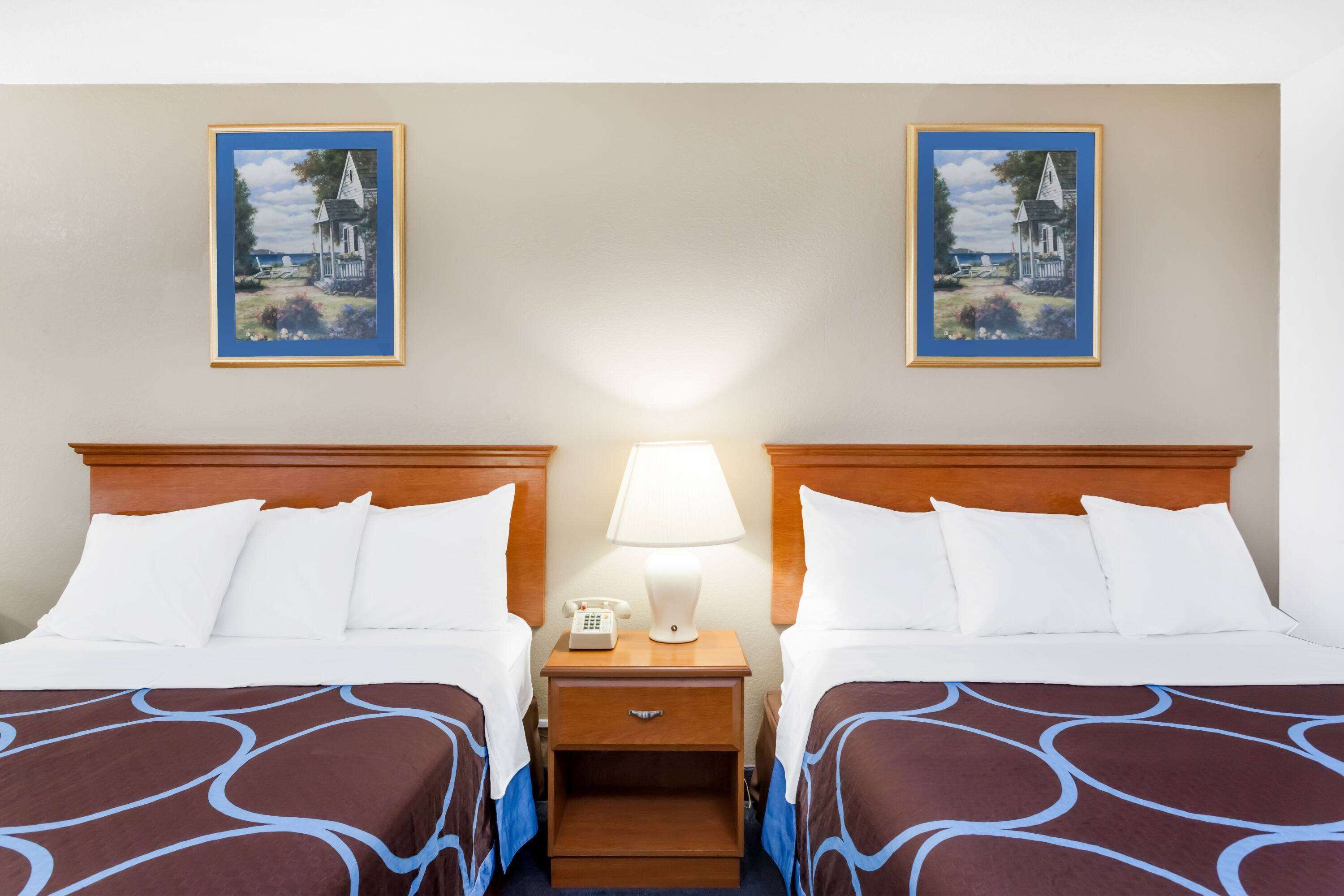 Habitación de hotel con dos camas, una mesita de noche con lámpara y dos cuadros en la pared.
