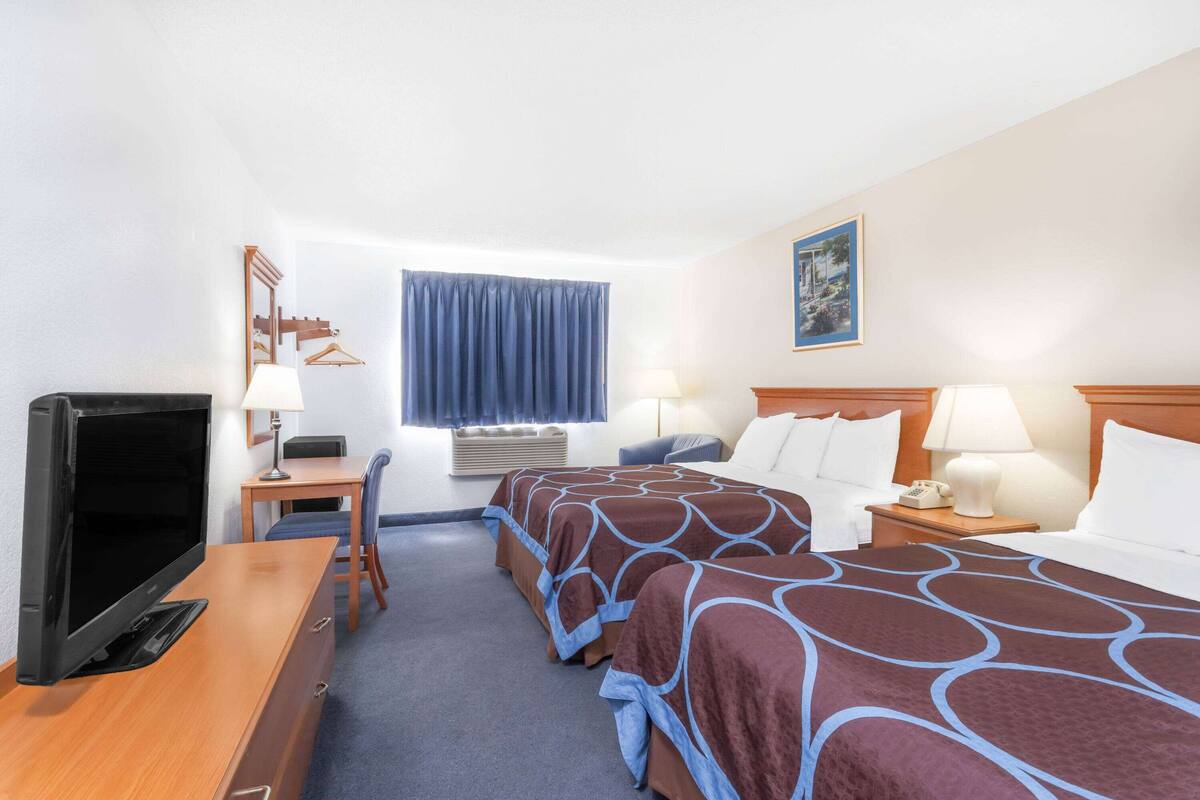 A hotel room with two beds, a desk with a TV, a chair, a desk lamp, a bedside table with a lamp, a window with curtains, and a painting on the wall.