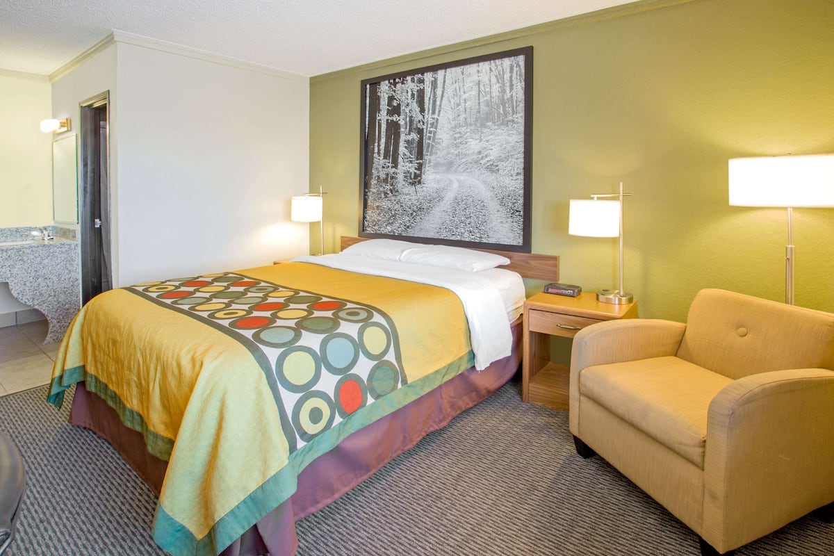 Quarto de hotel com cama, cadeira, mesa de cabeceira, luminária e um quadro na parede.