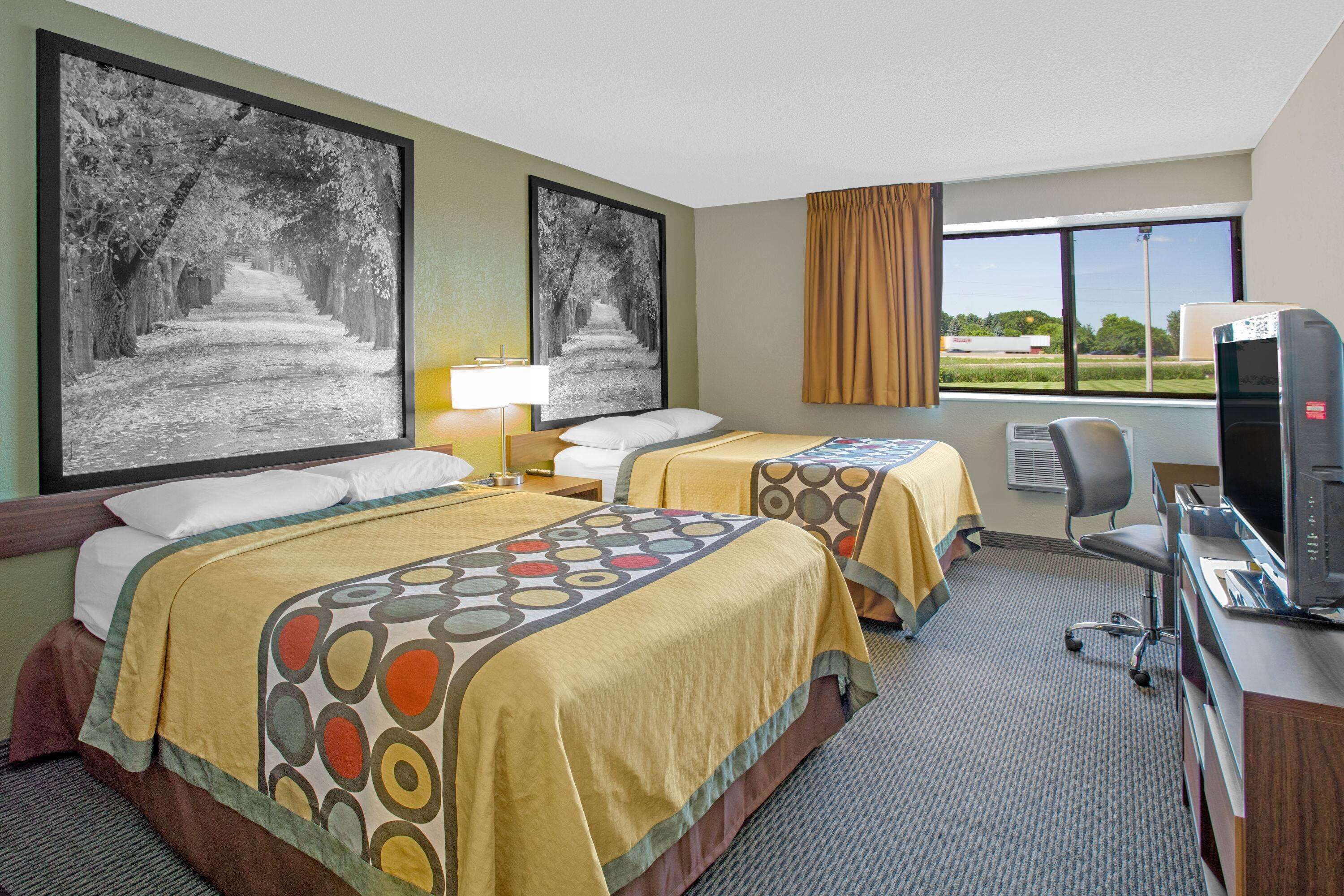 Een hotelkamer met twee bedden, een bureau met een computer, een televisie en grote ramen met uitzicht op het omliggende landschap.