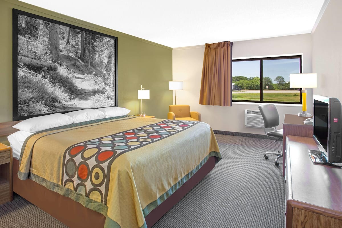 Quarto de hotel com uma cama grande, duas luminárias de cabeceira, uma escrivaninha com um computador e uma janela ampla com vista para o verde.