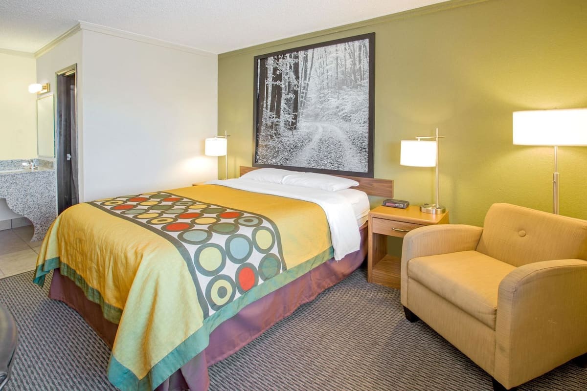 Quarto de hotel com uma cama, uma cadeira, uma escrivaninha e um quadro na parede.