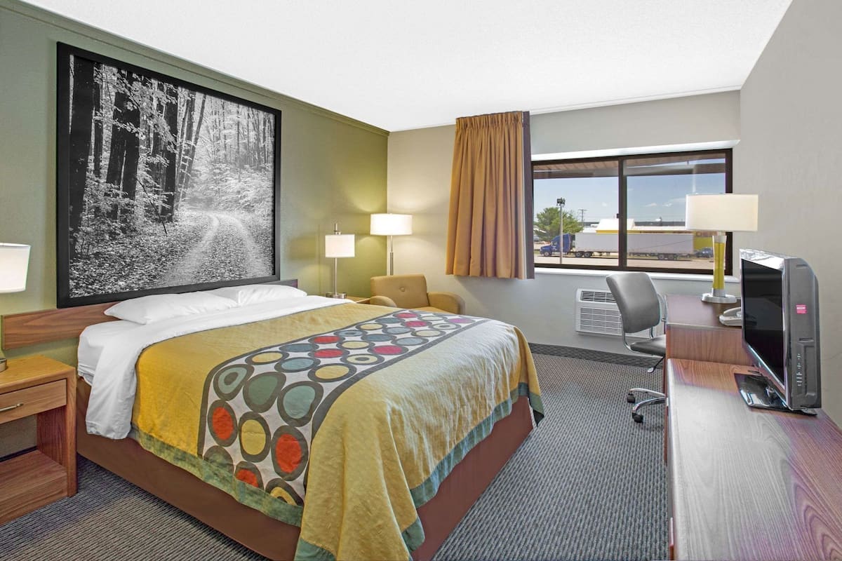 Quarto de hotel com uma cama, duas luminárias de cabeceira, uma escrivaninha com um computador, uma janela grande e um quadro na parede.
