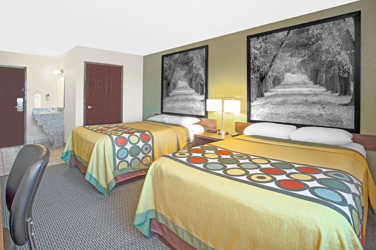 Quarto de hotel com duas camas, uma escrivaninha, uma cadeira, banheiro e duas grandes imagens emolduradas na parede.