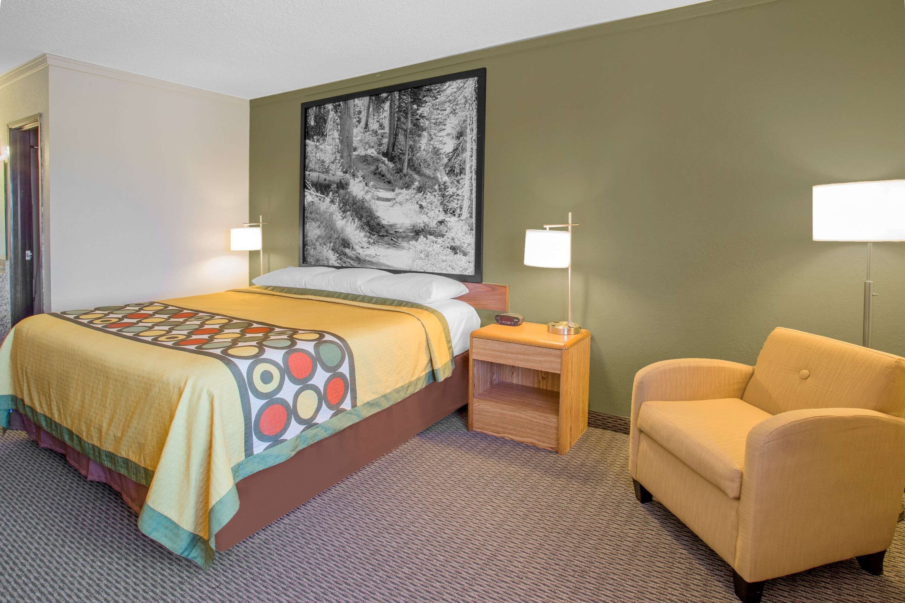 A hotel room with a bed, a chair, a nightstand, and a large framed picture on the wall.