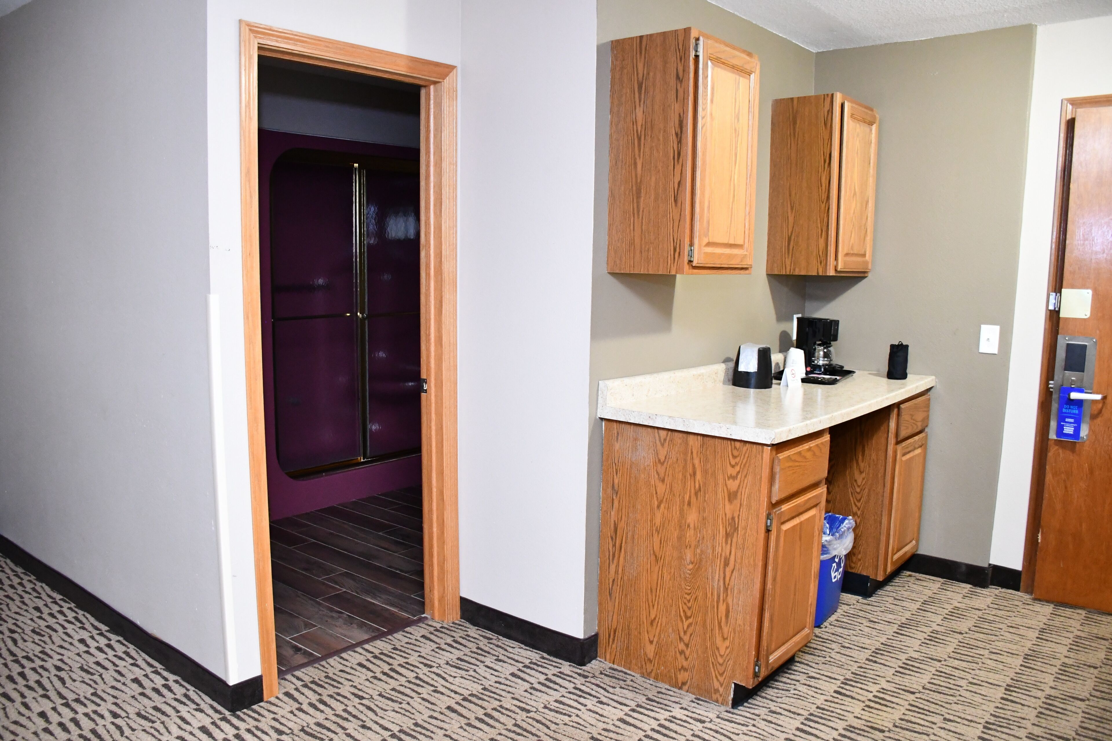 A hotel room with a kitchenette, a door leading to a purple room, and a patterned carpet.