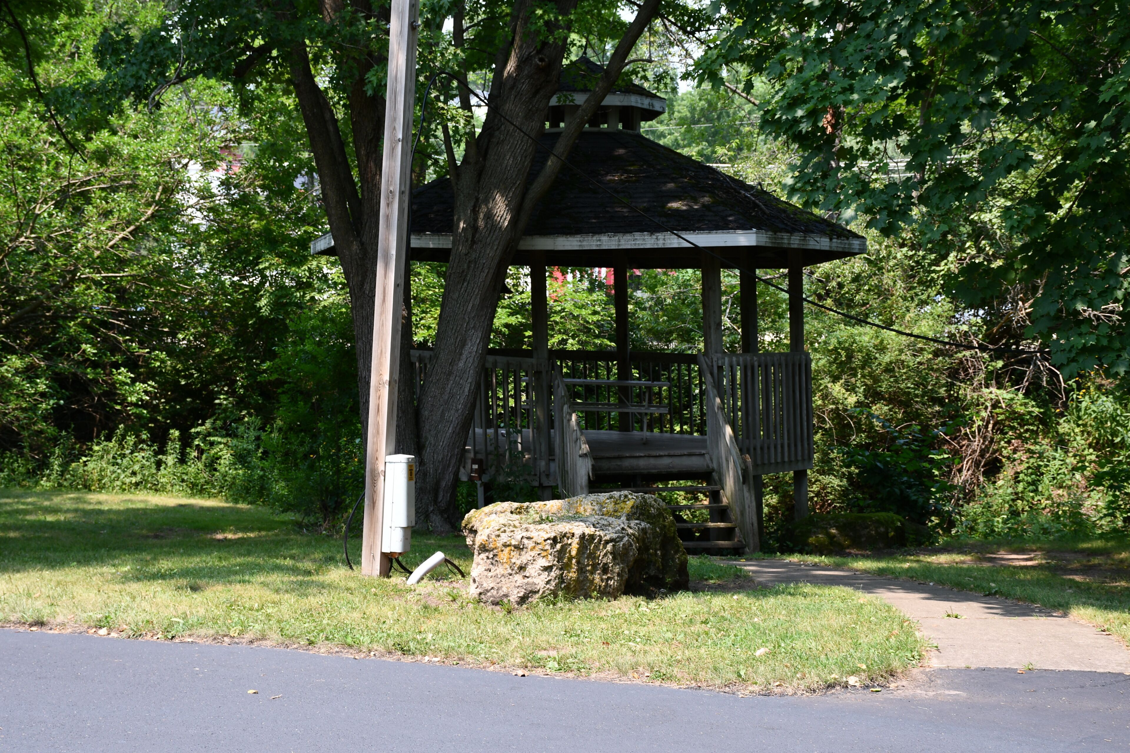 Ein Gartenpavillon mit Holzkonstruktion und Dach, umgeben von Bäumen und Gras.