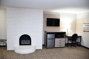A hotel room with a white brick fireplace, a flat-screen TV mounted on the wall, a desk with a lamp, a microwave, and a refrigerator.