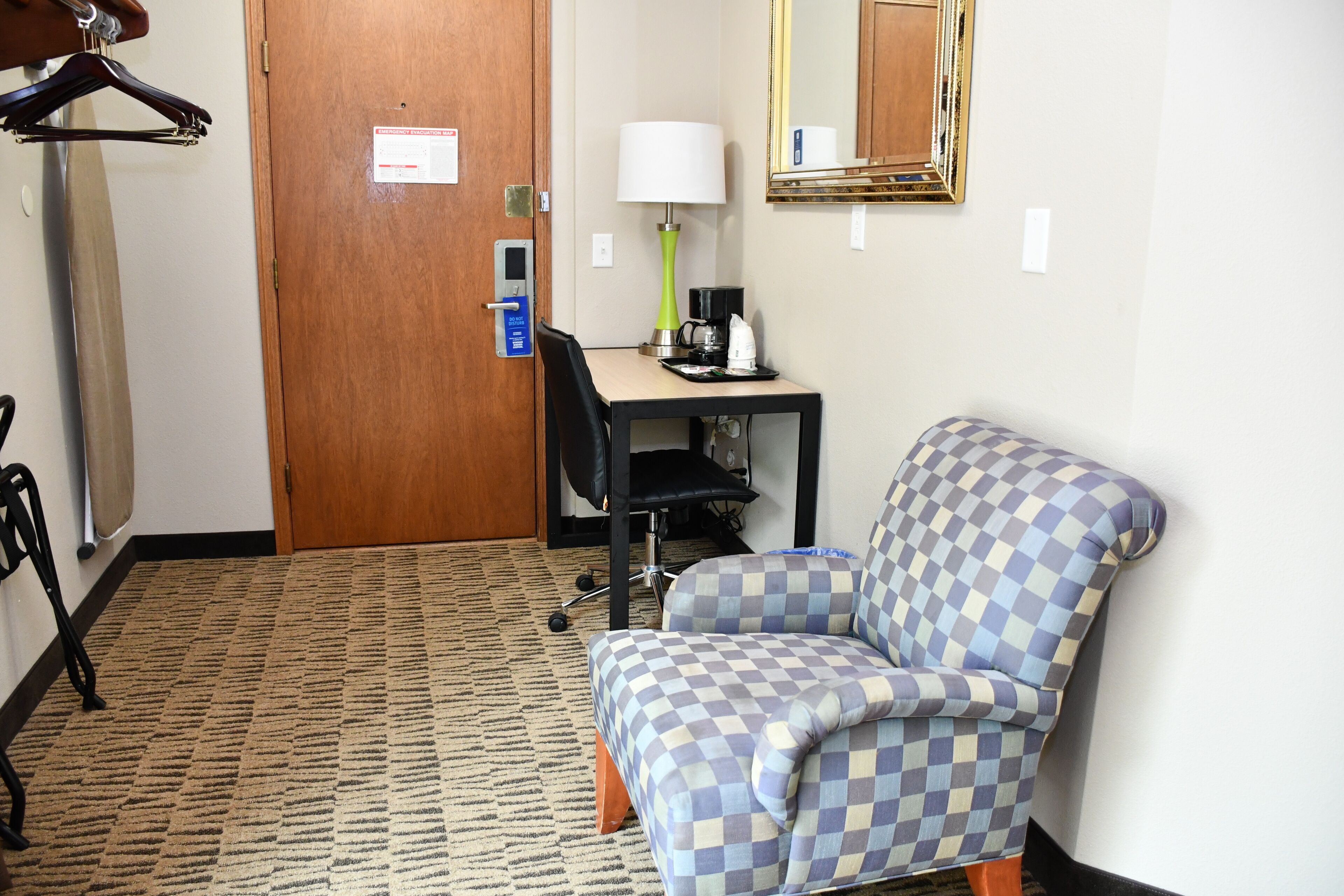 A hotel room with a checkered armchair, a desk with a lamp, a coffee maker, and a door with an emergency evacuation map.