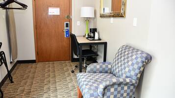 A hotel room with a checkered armchair, a desk with a lamp, a coffee maker, and a door with an emergency evacuation map.