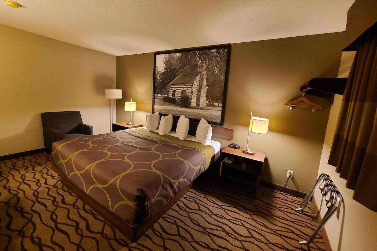 Une chambre d’hôtel avec un grand lit, deux tables de chevet, une chaise, un porte-manteau et un tableau accroché au mur.