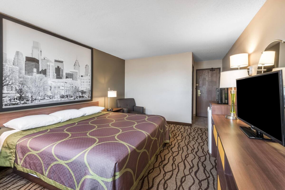 Une chambre d’hôtel avec un grand lit, une télévision, un bureau avec une lampe et un tableau représentant un paysage urbain accroché au mur.