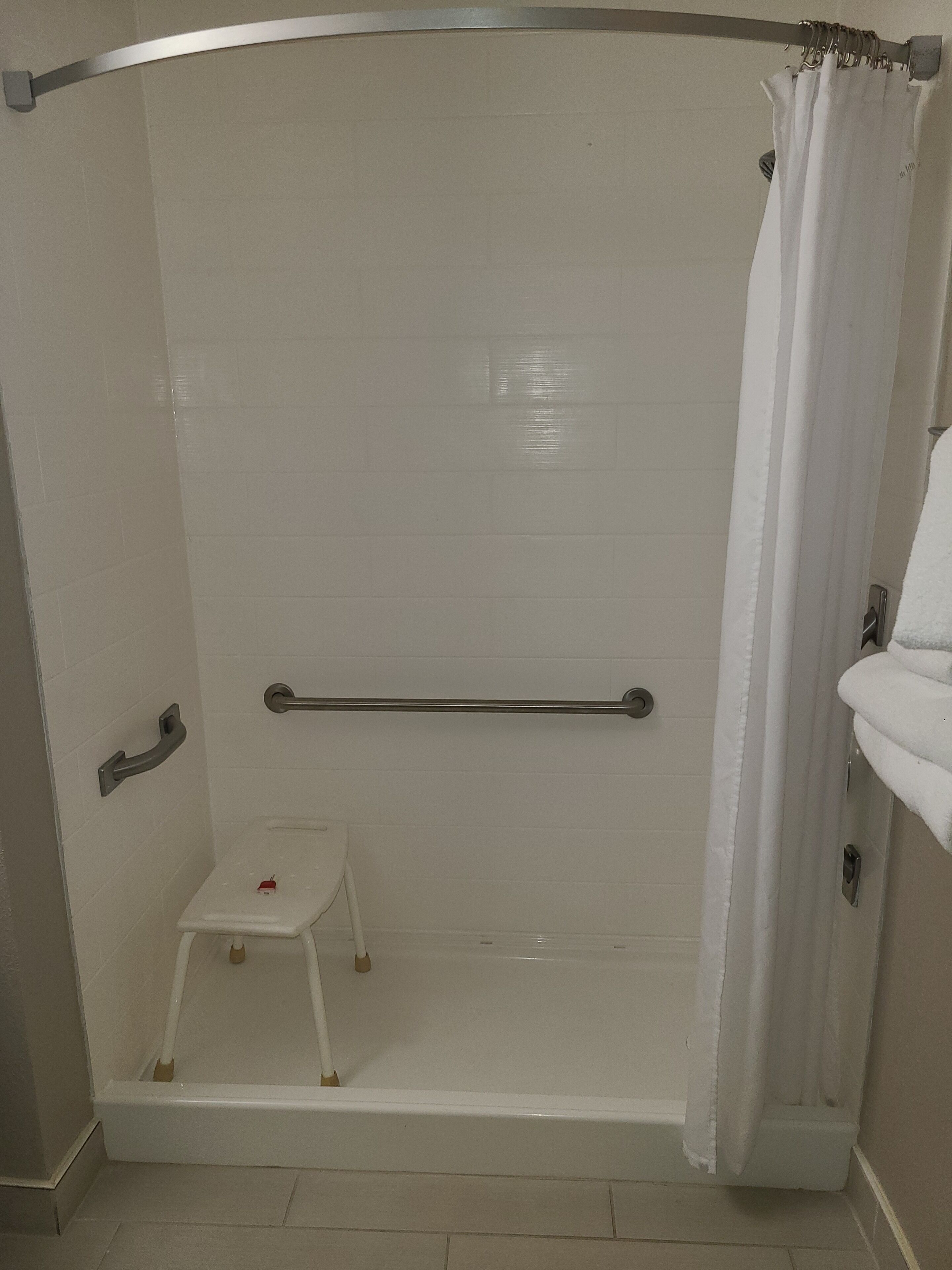 Une douche à l’italienne avec un mur recouvert de carreaux blancs, une barre d’appui, un petit marchepied et un rideau de douche.