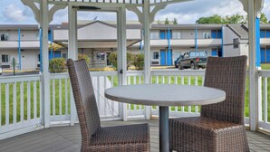 A covered outdoor area with wicker furniture and a white table, overlooking a parking area and buildings with blue doors. - Motel 6 - Harrisburg, PA - Near PA Expo Center  (Harrisburg)