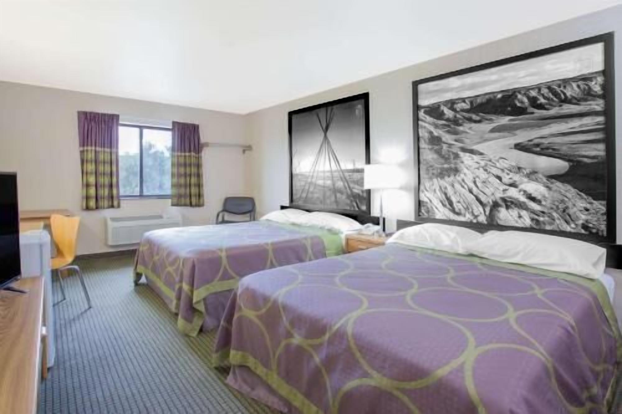 Une chambre d’hôtel avec deux lits, un bureau, une chaise, une fenêtre avec des rideaux et un grand tableau représentant un paysage en noir et blanc accroché au mur.
