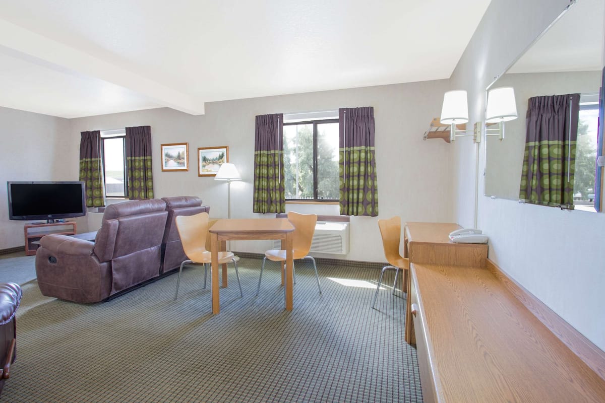 A hotel room with a sofa, a TV, a dining table with chairs, a small kitchen area, and a large window with curtains.