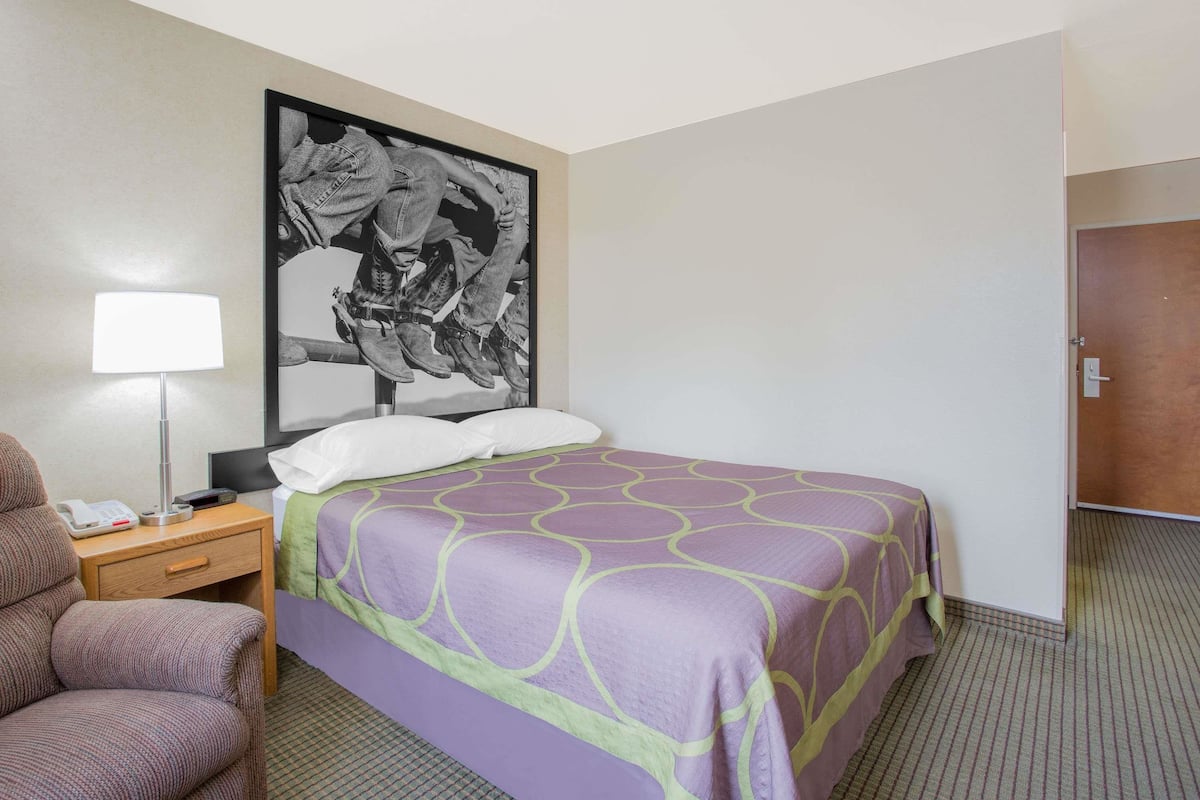 A hotel room with a bed, a chair, a nightstand with a lamp, and a door.
