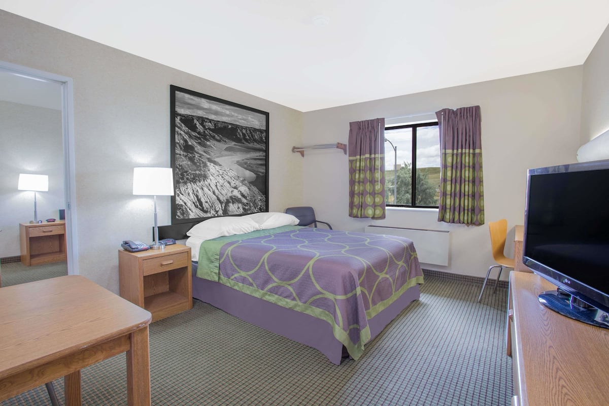 A hotel room with a bed, a desk, a TV, a window with curtains, and a painting on the wall.