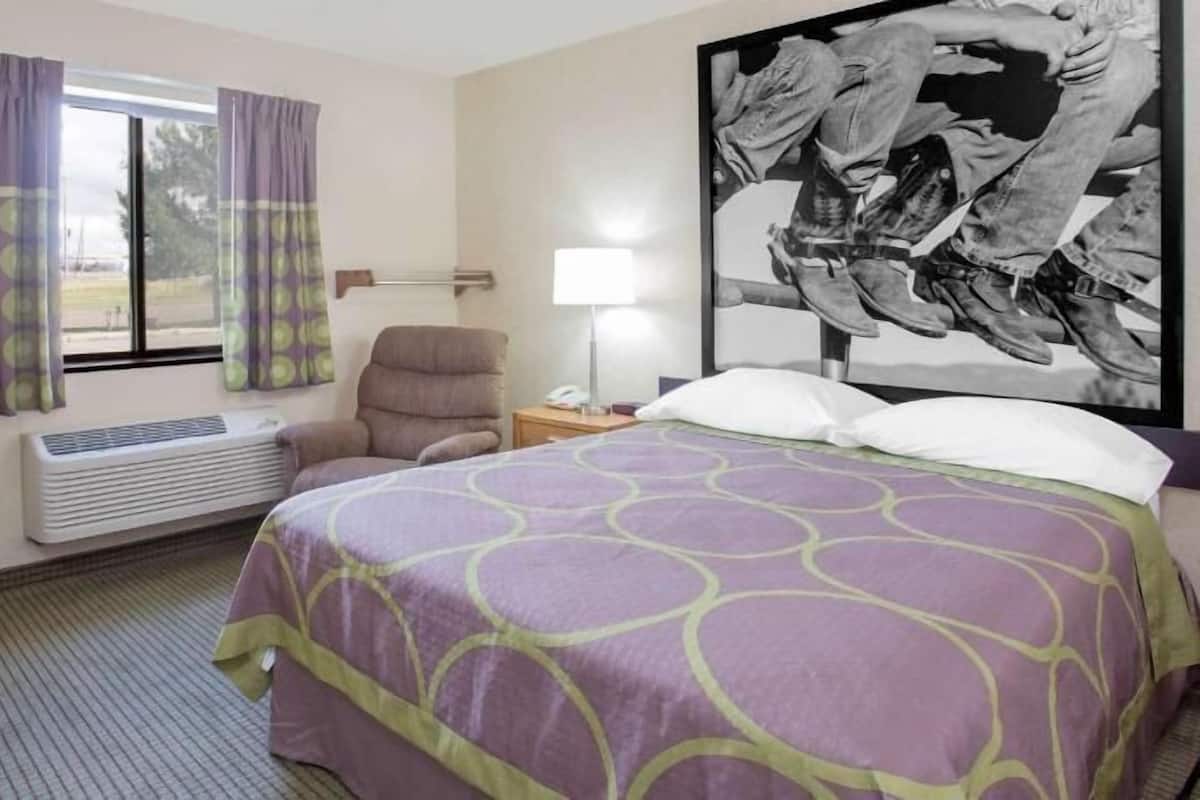 A hotel room with a bed, a chair, a desk, a lamp, a window with curtains, and a wall-mounted air conditioner.