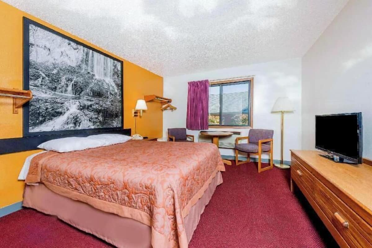 Quarto de hotel com cama grande, escrivaninha com cadeira, televisão e janela com cortinas.