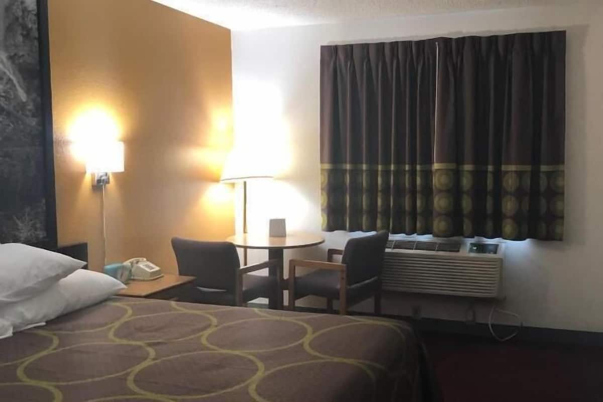 Quarto de hotel com cama, escrivaninha com cadeira, luminária, janela com cortinas e televisão.