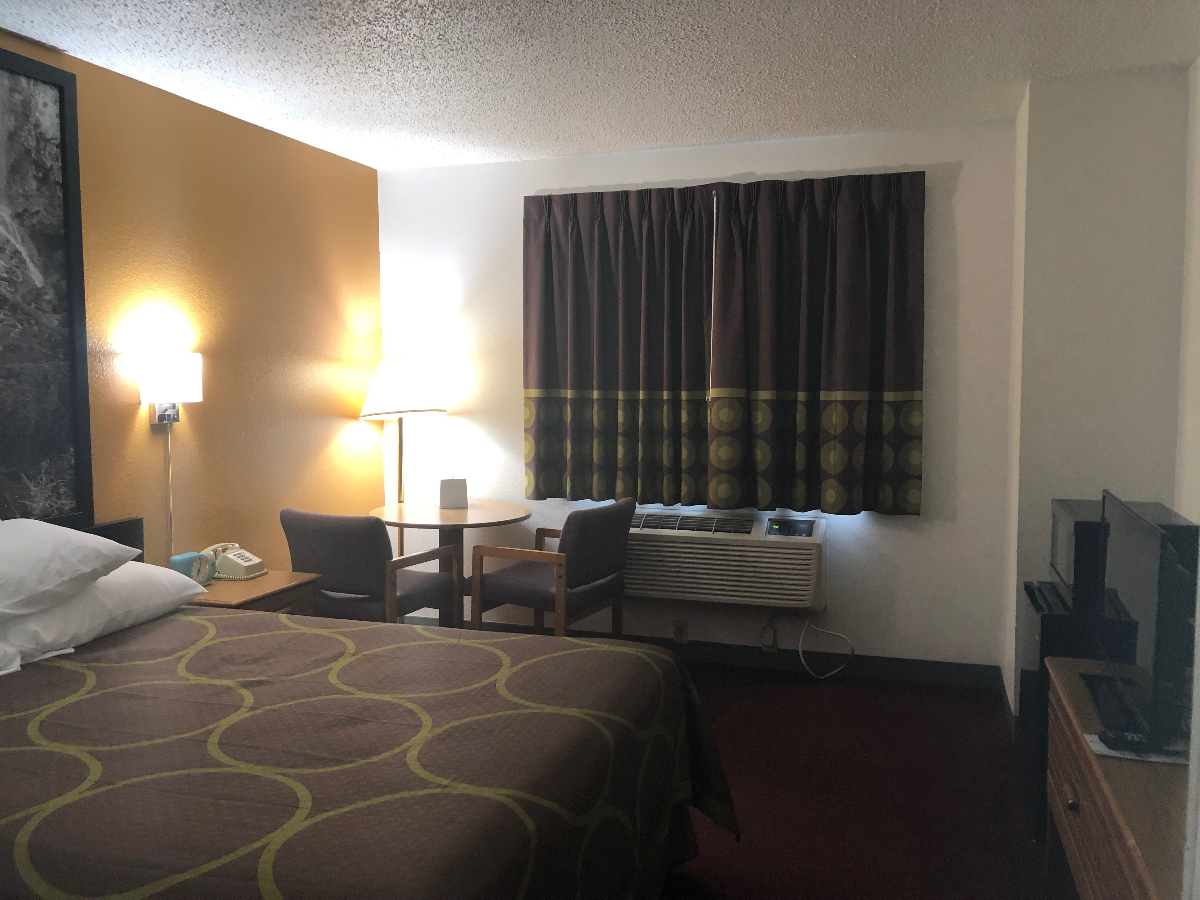 A hotel room with a bed, a desk with a chair, a lamp, an air conditioning unit, and a television.