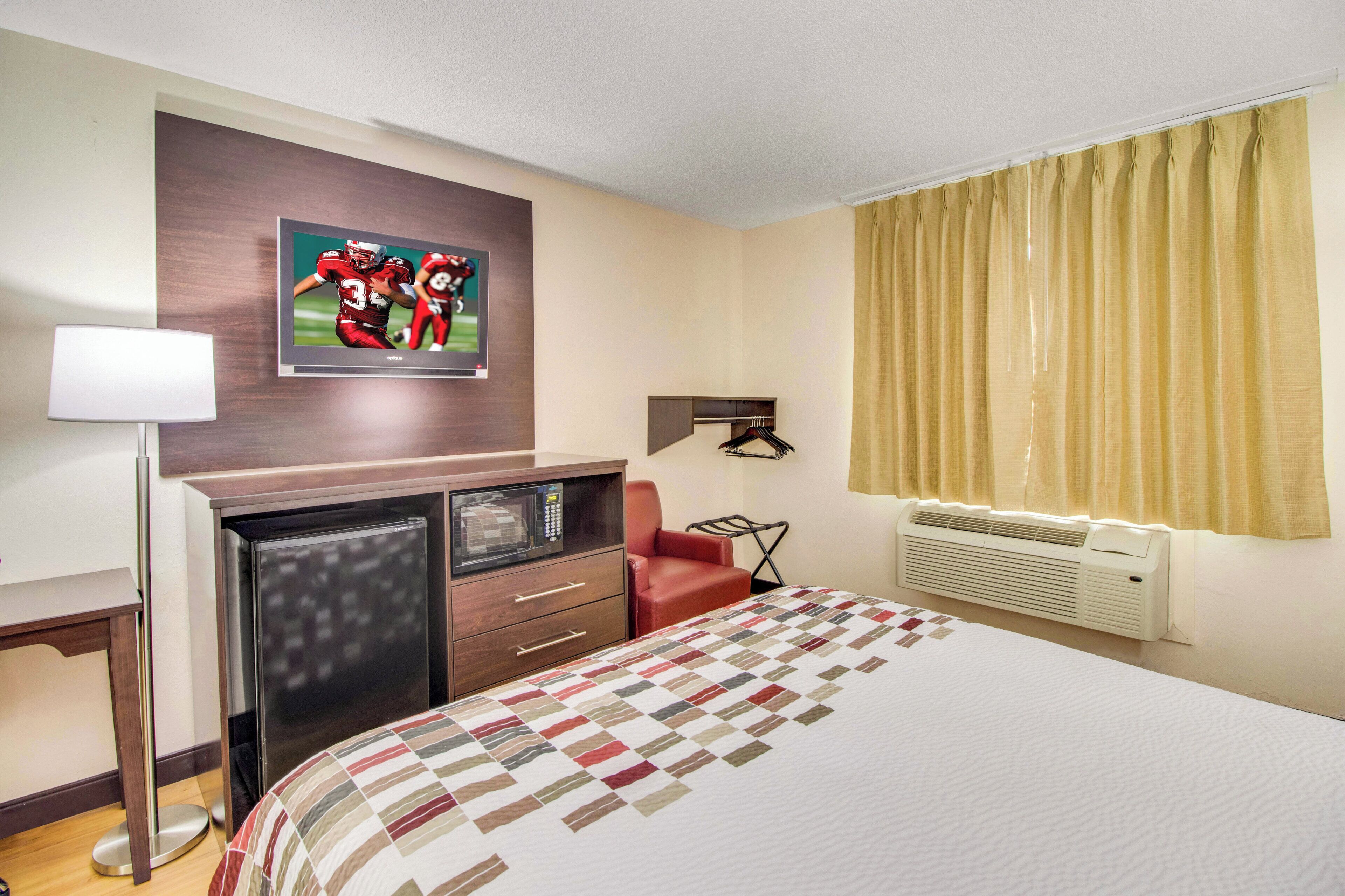 Une chambre d’hôtel avec un lit, un bureau doté d’un micro-ondes, une chaise rouge, un téléviseur fixé au mur et une fenêtre avec des rideaux.
