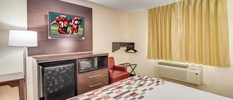 A hotel room with a bed, a desk with a microwave, a red chair, a wall-mounted TV, and a window with curtains.