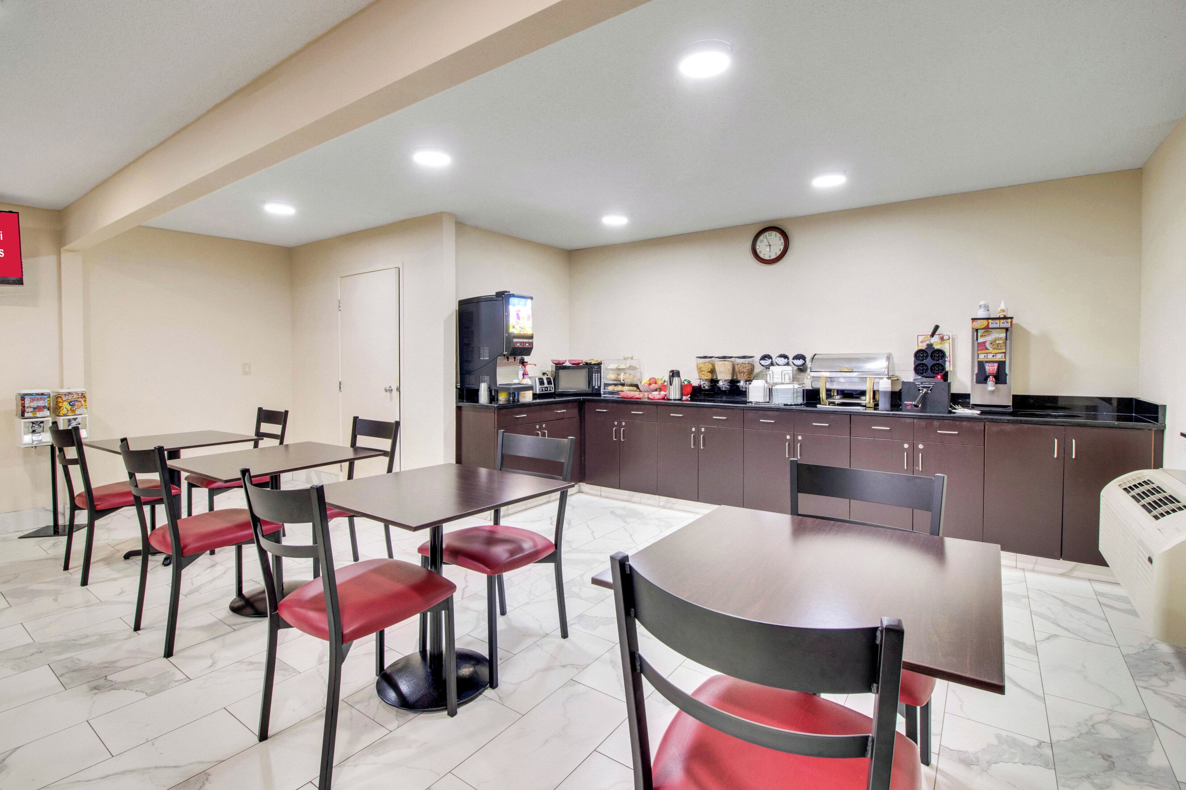 A dining area with tables and chairs, a kitchen with appliances, and a clock on the wall.