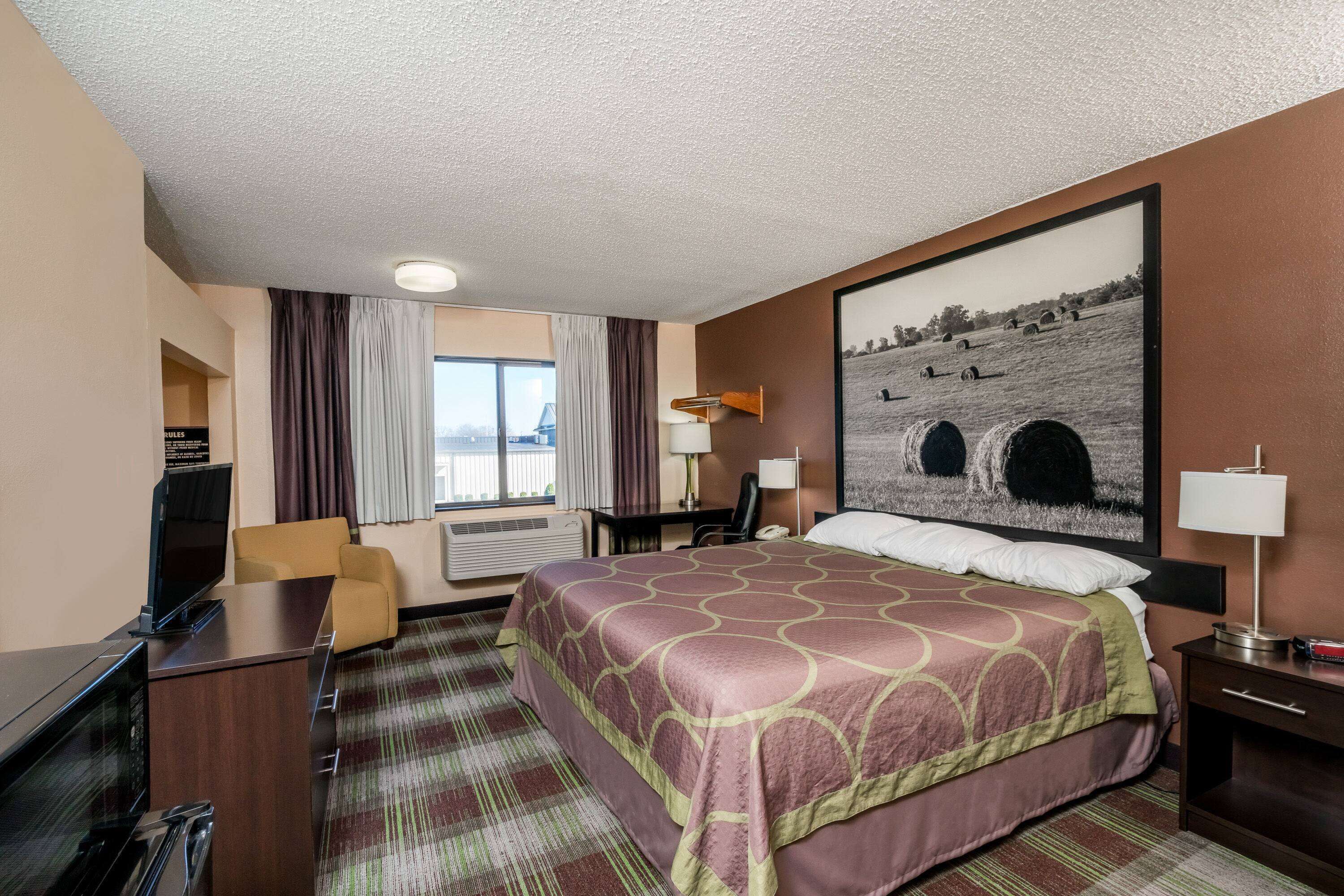 A hotel room with a large bed, a desk with a television, a chair, a desk lamp, and a painting of a landscape on the wall.