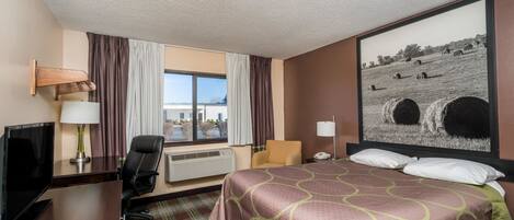 Habitación de hotel con cama, escritorio con televisor, una silla, ventana con cortinas y un cuadro en la pared.