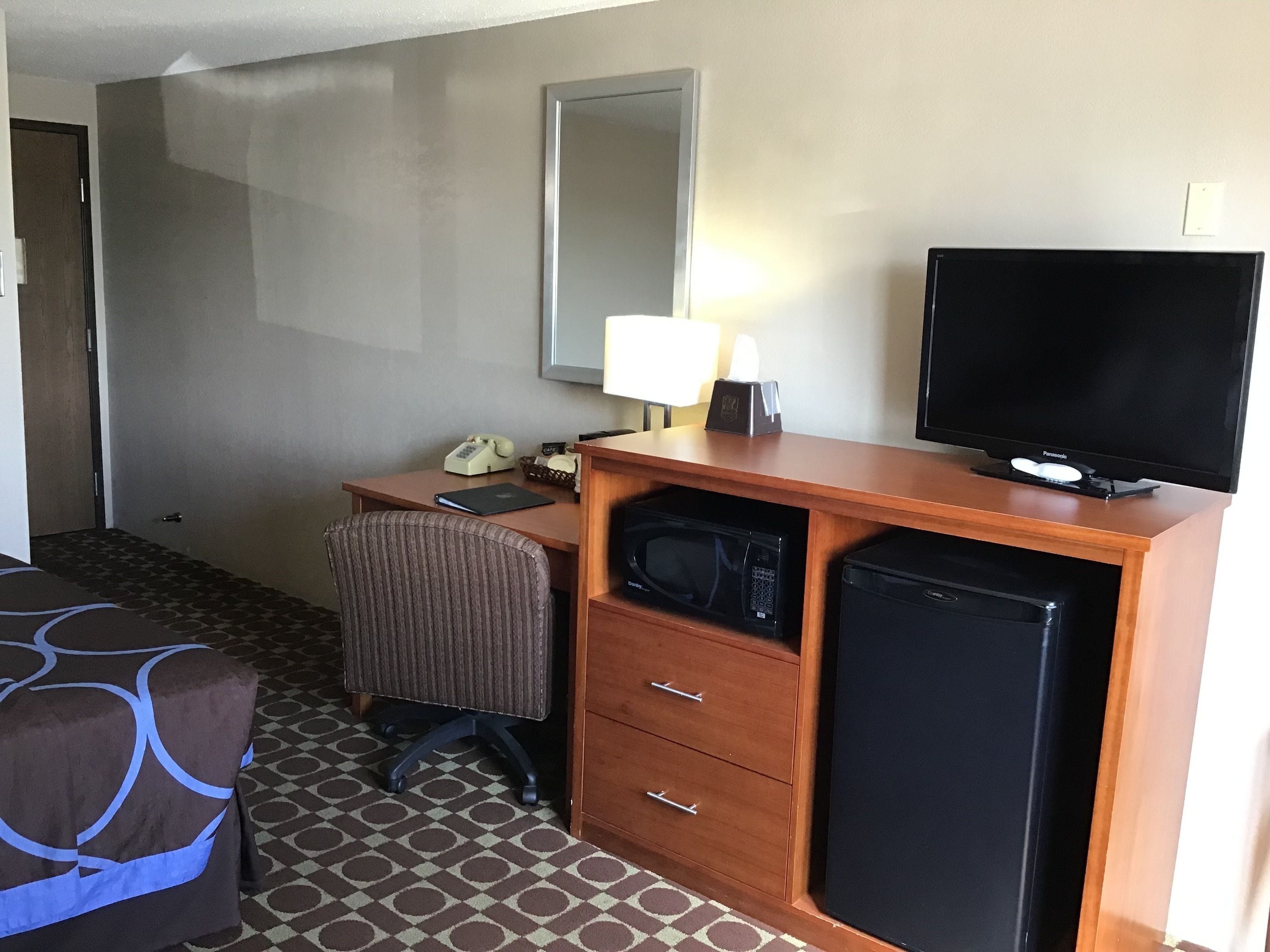 A hotel room with a wooden desk, a television, a microwave, a refrigerator, a chair, and a bed with a patterned cover.