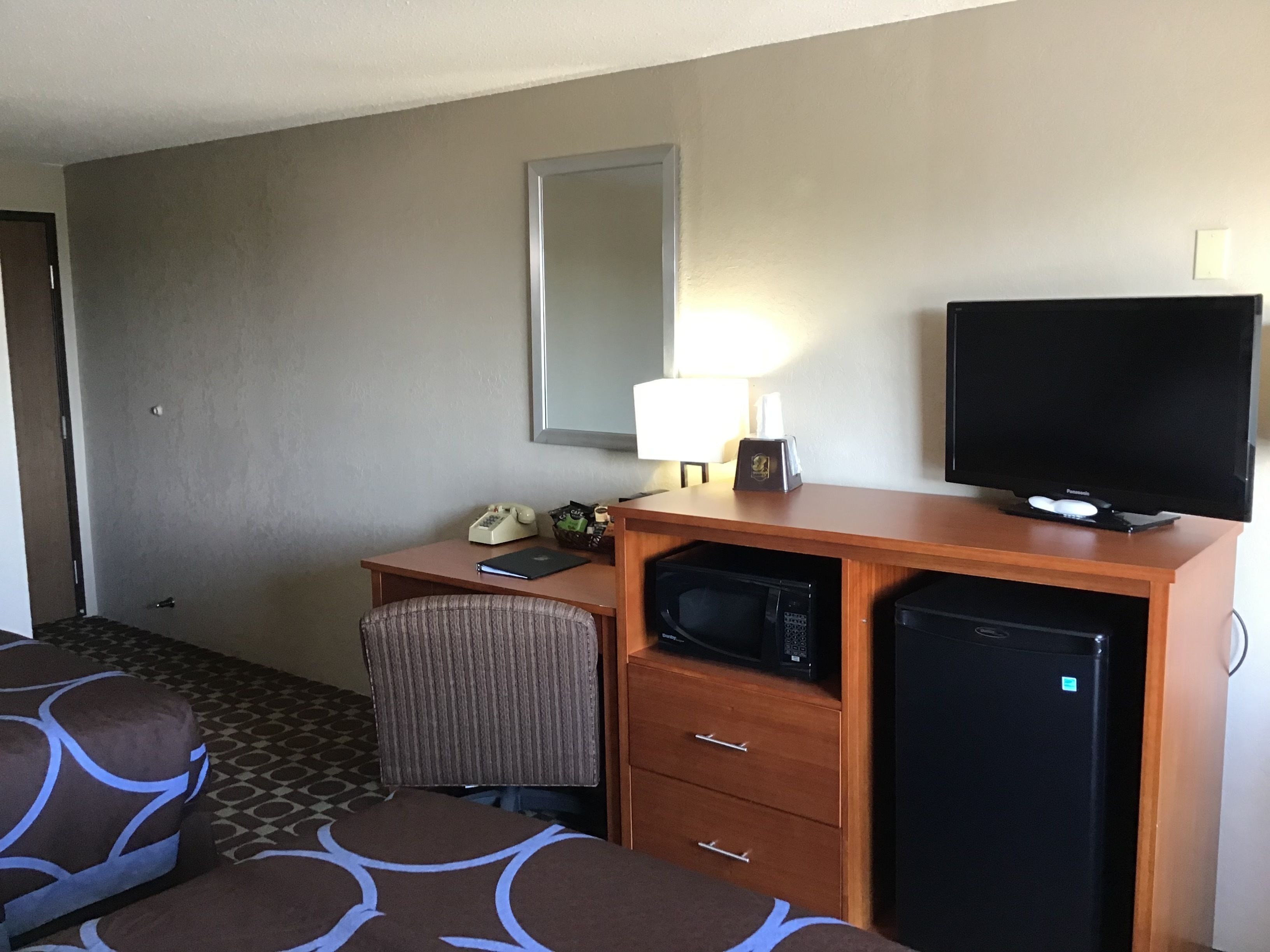 A hotel room with a wooden desk, a television, a microwave, a refrigerator, a chair, and a bed with patterned bedding.