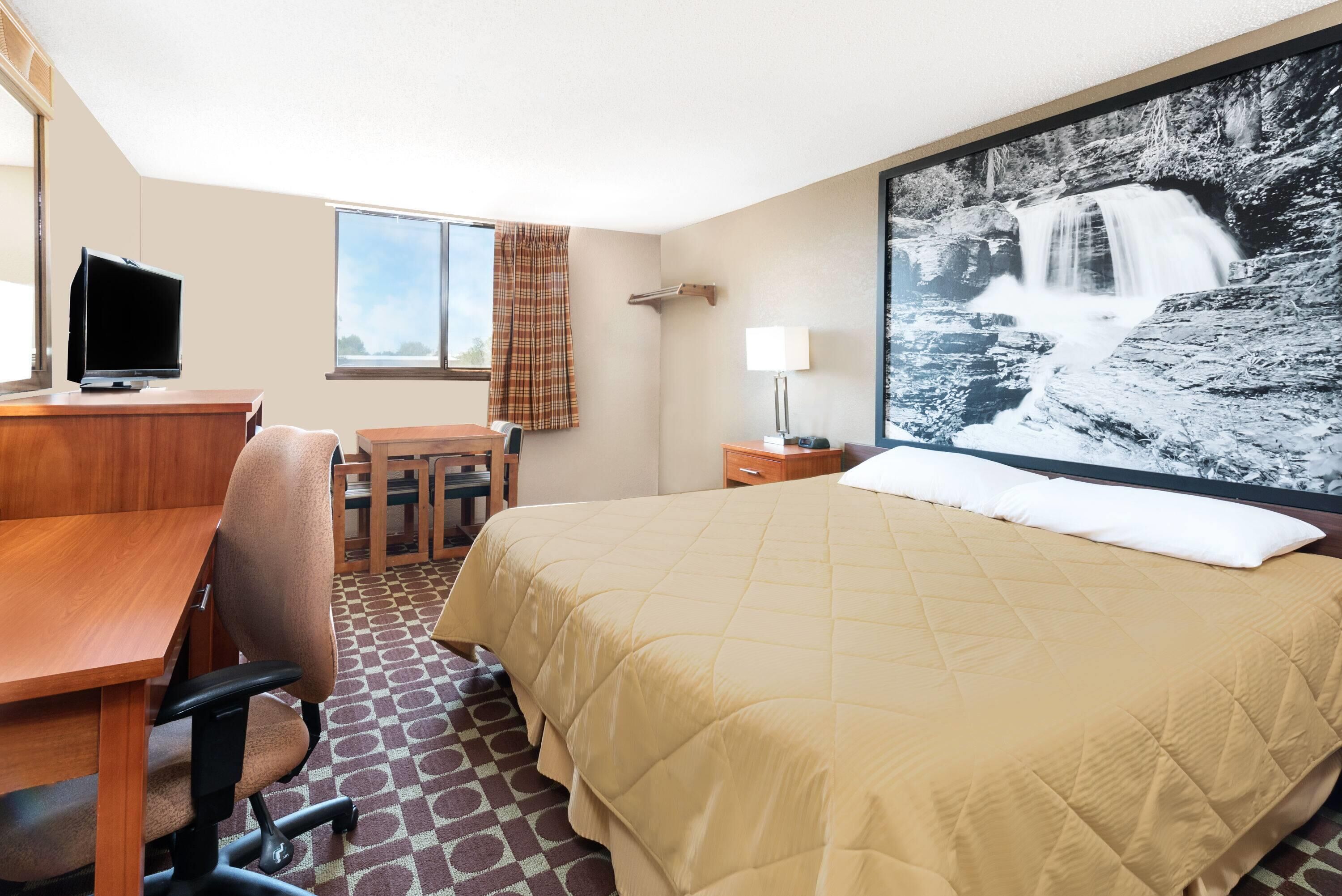 A hotel room with a bed, a desk with a chair, a television, a window with curtains, and a large wall-mounted picture of a waterfall.