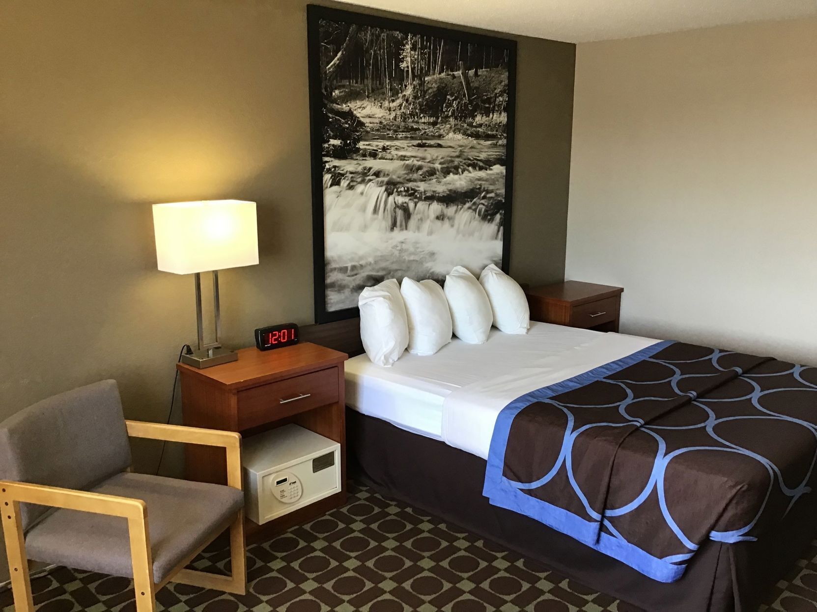 A hotel room with a bed, a desk with a lamp, a chair, a nightstand, and a painting of a waterfall.