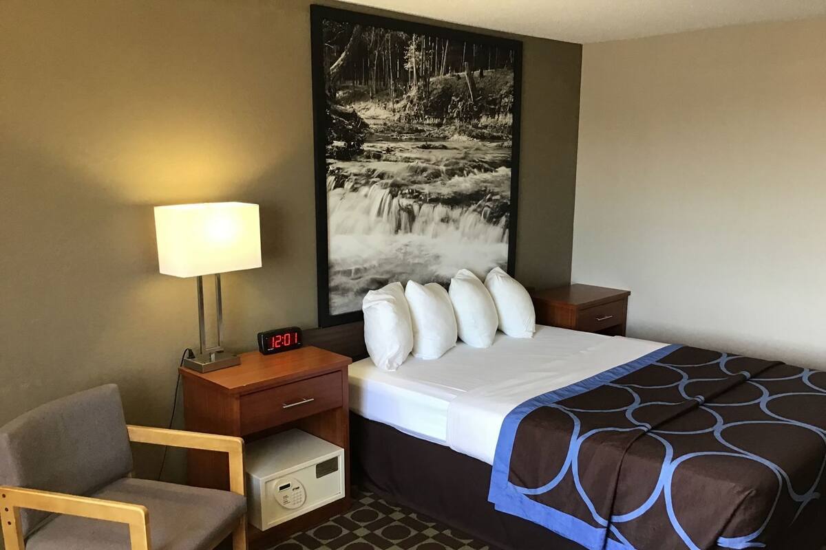A hotel room with a bed, a desk with a lamp, a chair, a nightstand, and a painting of a waterfall.